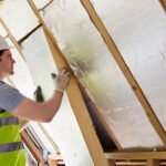 A construction worker in safety gear (hard hat and high-visibility vest) inspects insulation between wooden roof beams during a home renovation. They're wearing protective gloves and examining the white insulation material installed in the sloped ceiling space between exposed timber rafters.